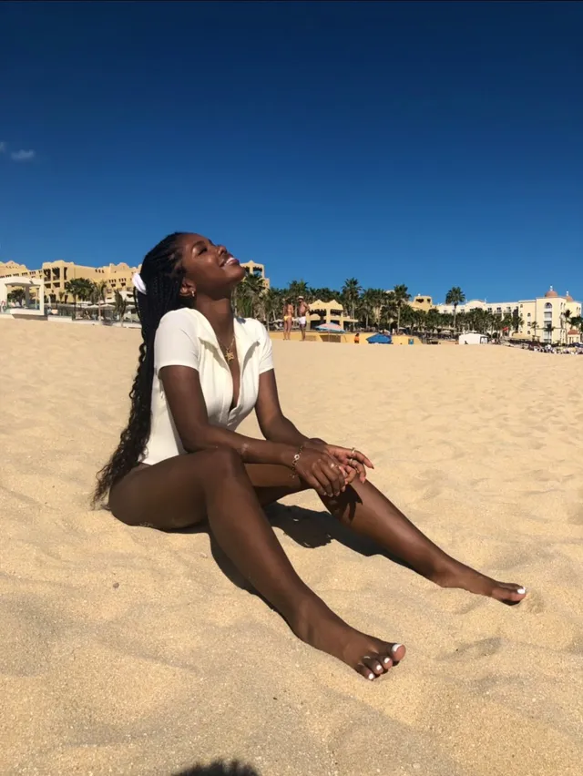 Ryan Destiny sitting barefoot on a sandy beach in a white top and shorts.