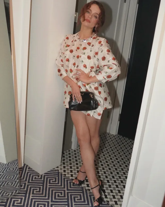 Joey King posing in a floral outfit and black heeled sandals.