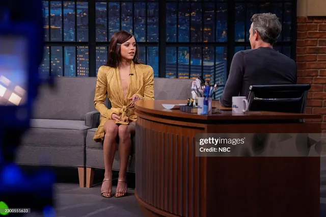 Jenna Ortega sitting during a television interview in a yellow outfit and heels.