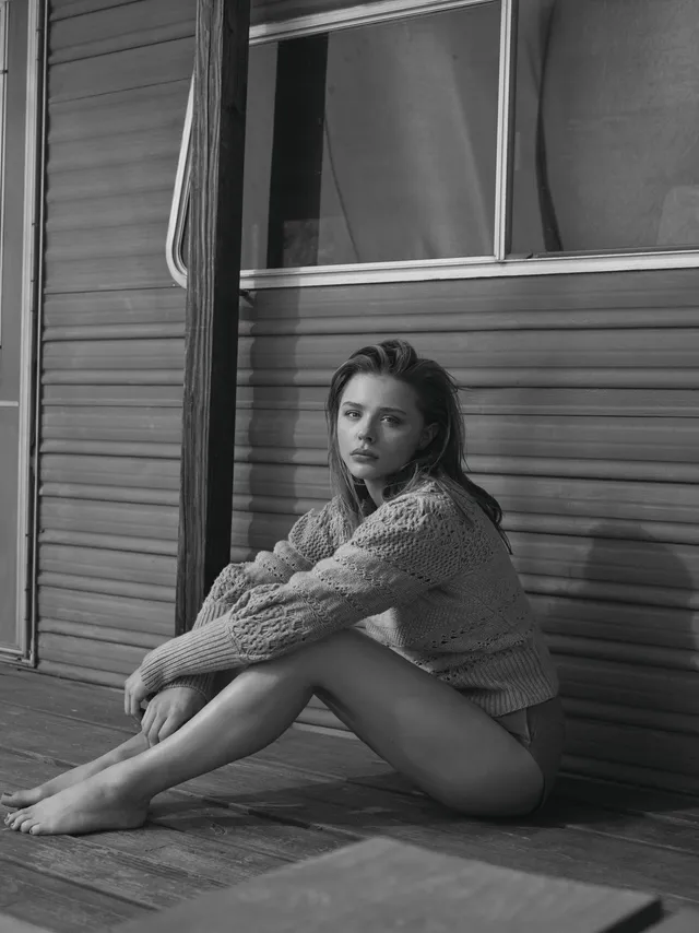 Chloë Grace Moretz sitting barefoot beside a window in a monochrome portrait.