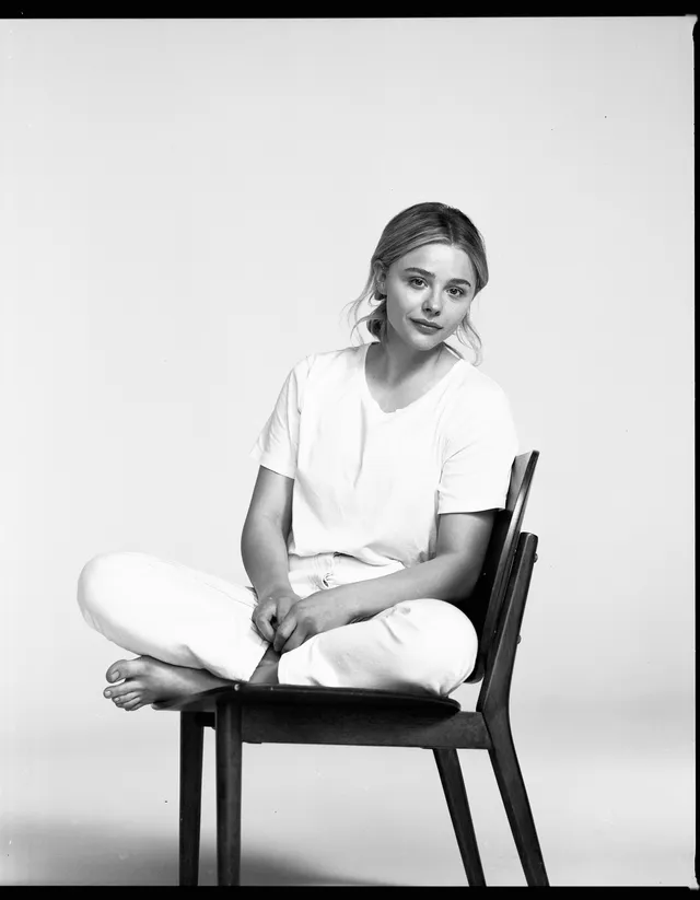 Chloë Grace Moretz sitting barefoot on a chair in a monochrome portrait.