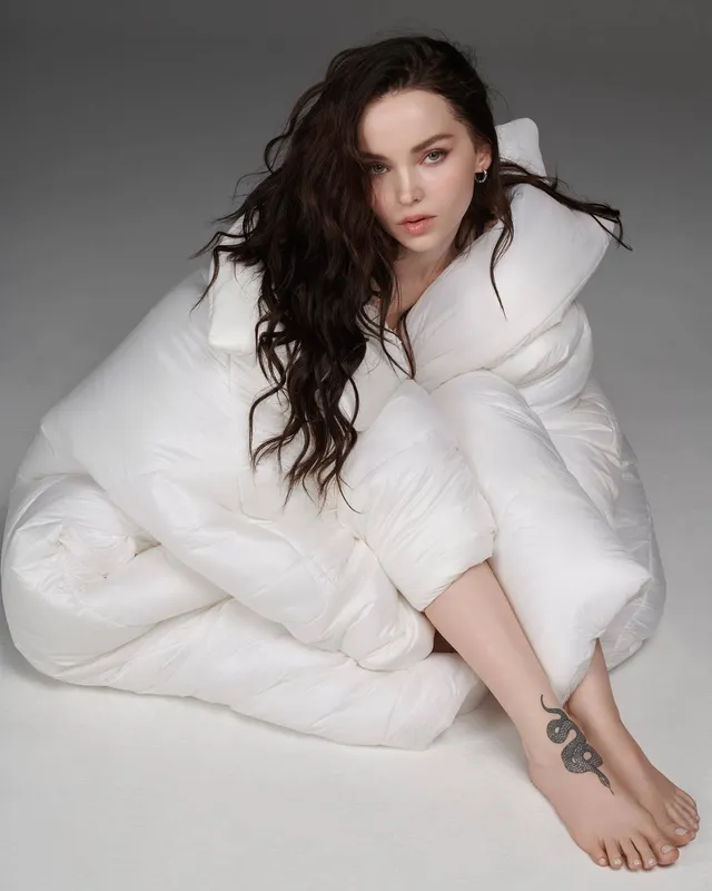 Dove Cameron sitting barefoot in a white studio outfit.