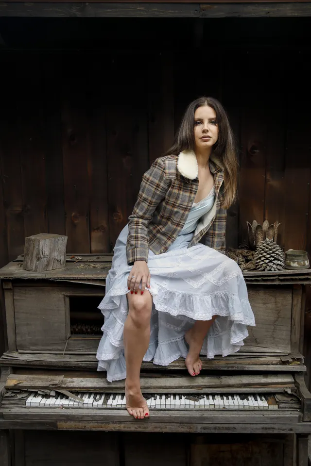 Lana Del Rey sitting barefoot on a piano in a layered pale skirt.