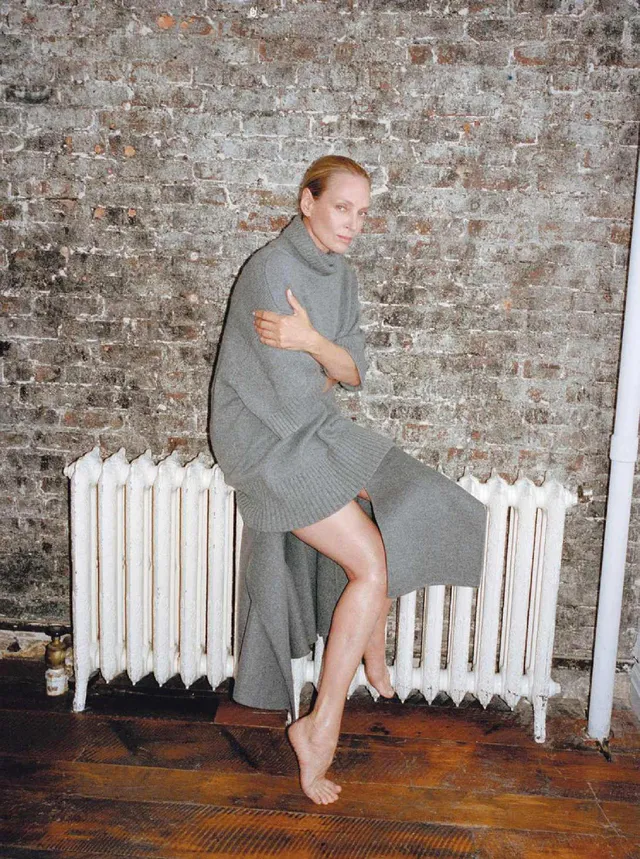 Uma Thurman sitting barefoot in a gray outfit against a brick wall.
