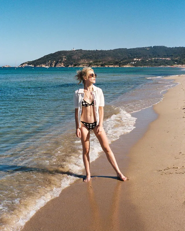 Kiernan Shipka standing barefoot at the water's edge on a beach.