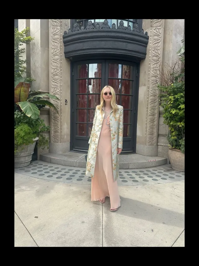 Dakota Fanning standing outdoors in a pale outfit and long coat.