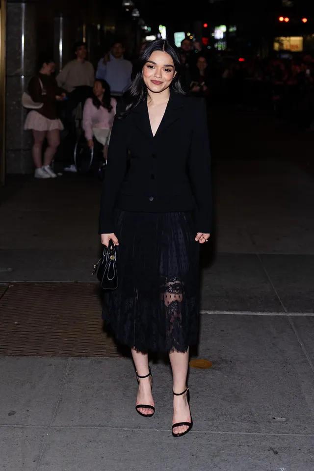 Rachel Zegler standing outside in a black dress and heeled sandals.