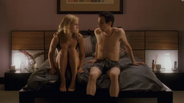 Alice Eve sitting on a bed beside a man in a screen scene.