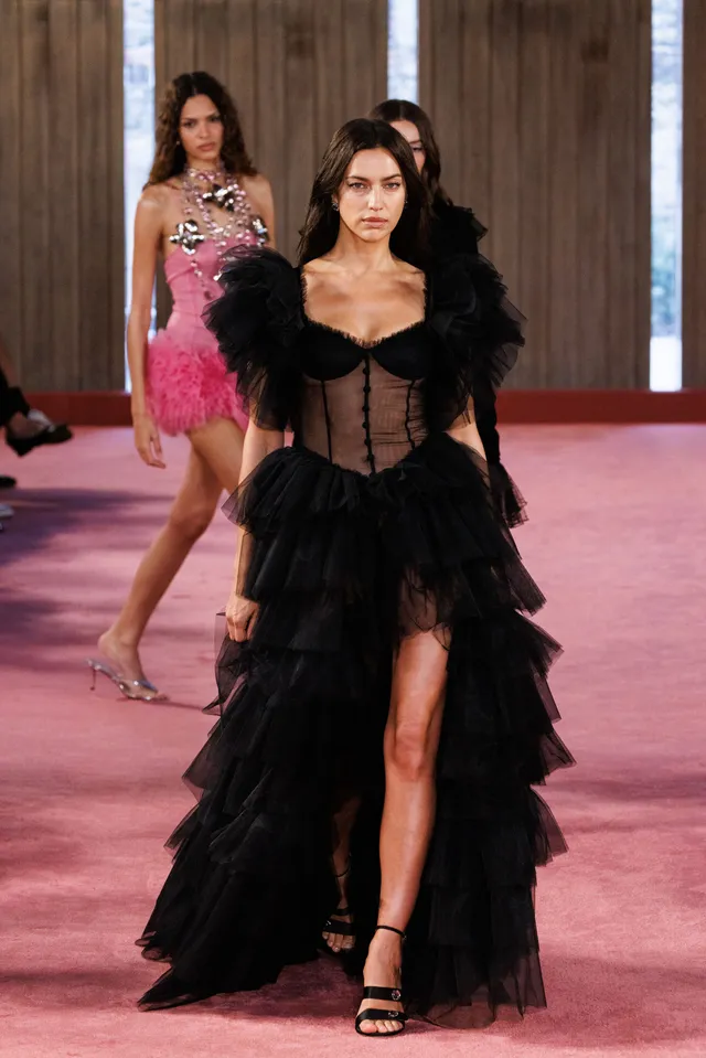 Irina Shayk walking a pink runway in a long black outfit and heels.