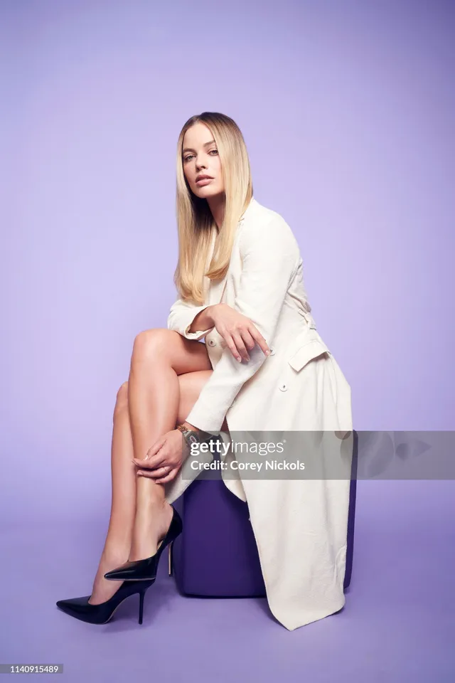 Margot Robbie sitting on a purple block in a white outfit and black pointed heels.