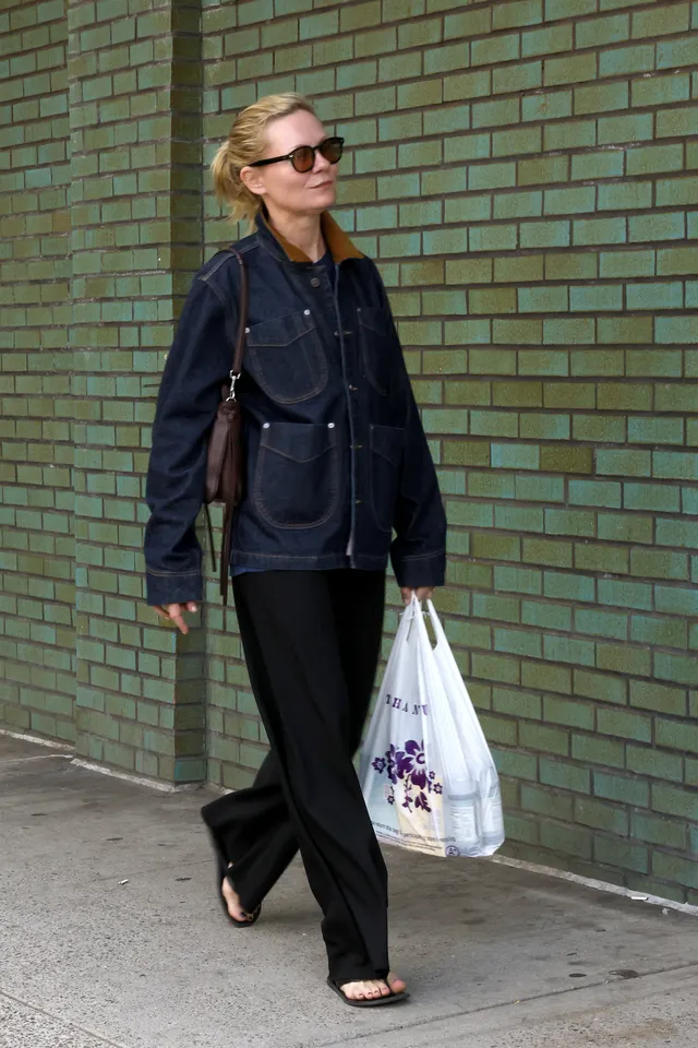 Kirsten Dunst walking beside a brick wall in a dark jacket and sandals.