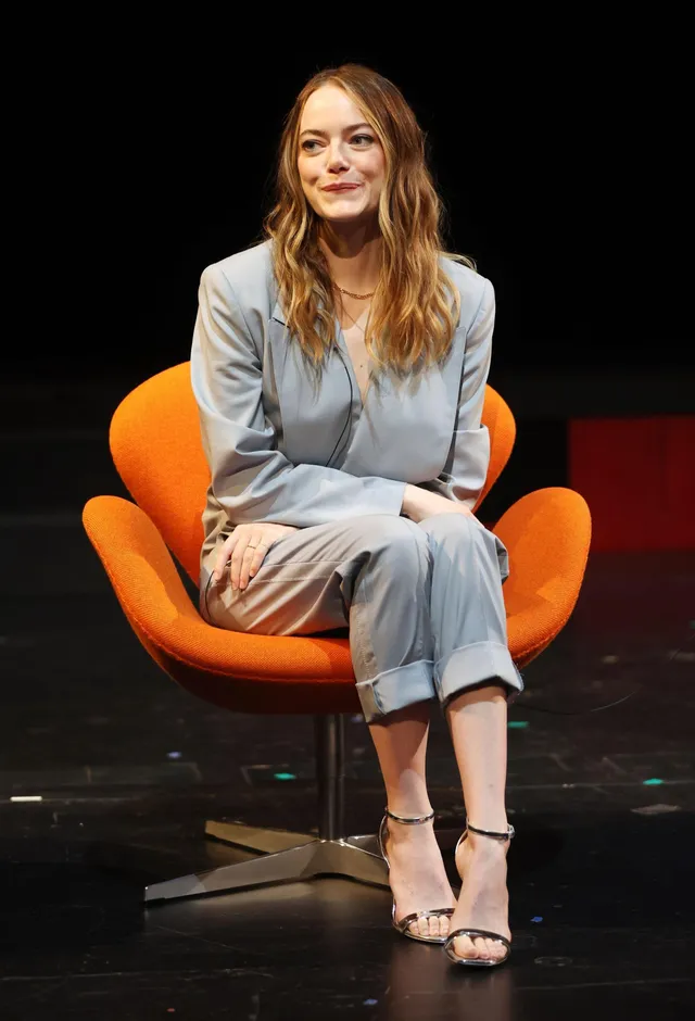 Emma Stone sitting in an orange chair in a pale suit and metallic ankle-strap heels.