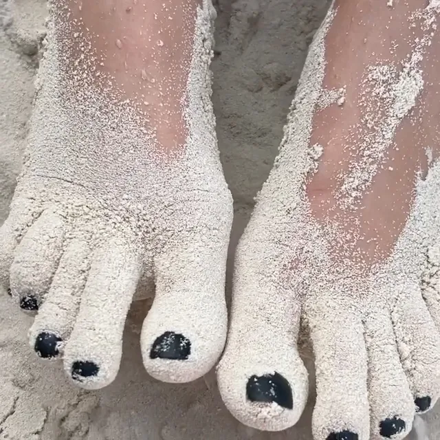 Olivia Cooke showing sand covered feet with dark nail polish.