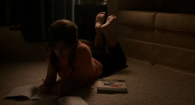 Cailee Spaeny lying barefoot on a carpet beside a bed in a film scene.