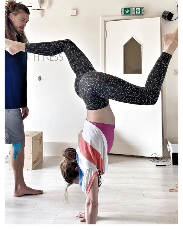 Rebecca Ferguson performing a handstand barefoot in a workout room beside a companion.