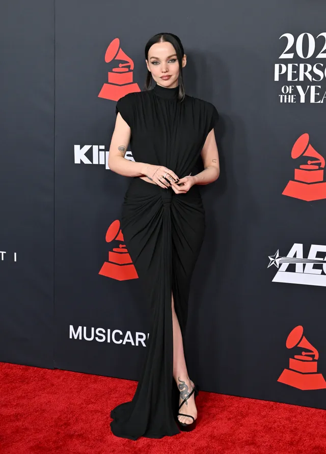 Dove Cameron wearing a black gown and strappy heels on a carpet.