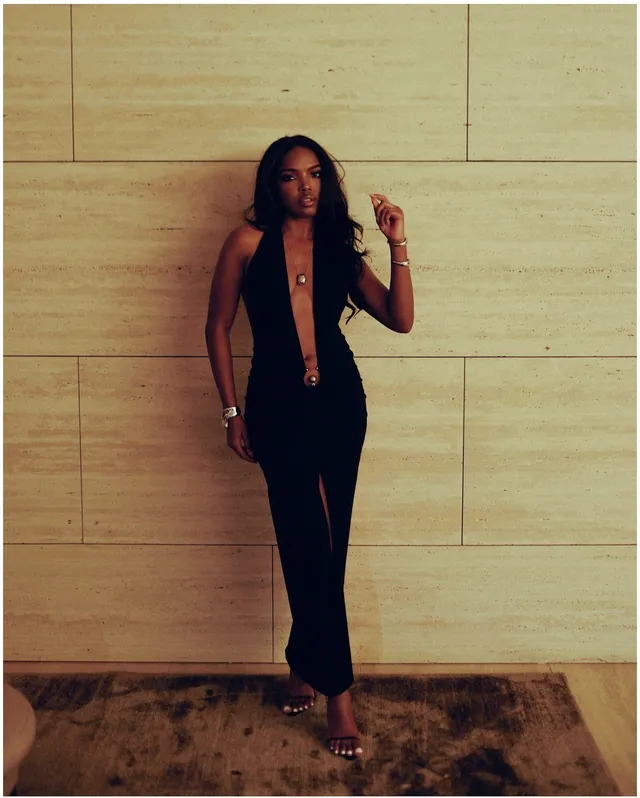 Ryan Destiny wearing a black halter dress and metallic heels against a pale wall.