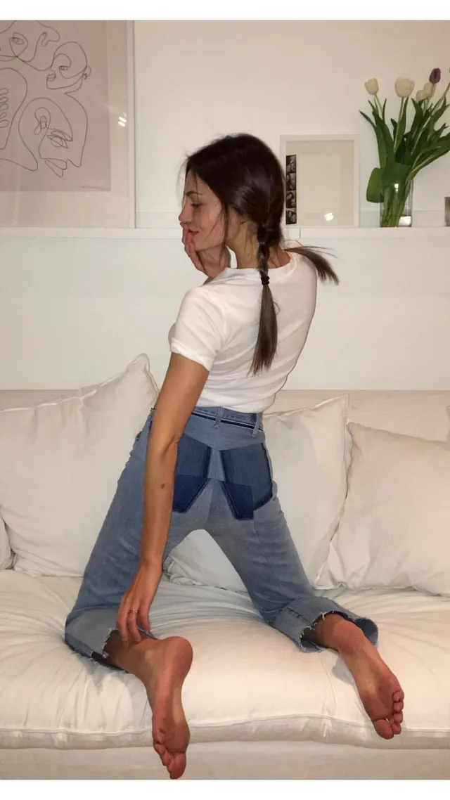 Phoebe Tonkin kneeling on a sofa in a white top and jeans.