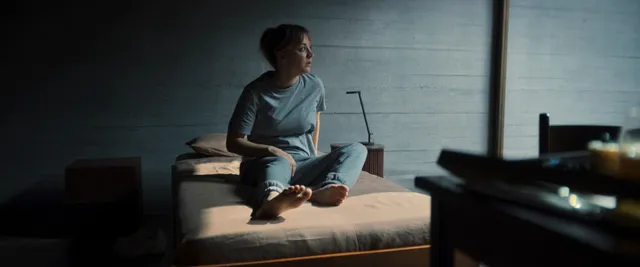 Kaley Cuoco sitting barefoot on a bed in a dimly lit film scene.