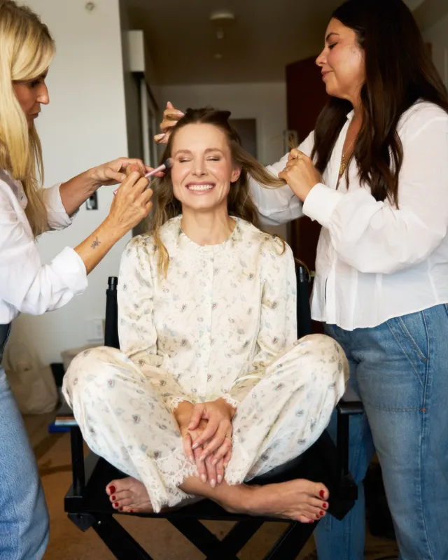 Kristen Bell sitting barefoot in a makeup chair while two people assist.