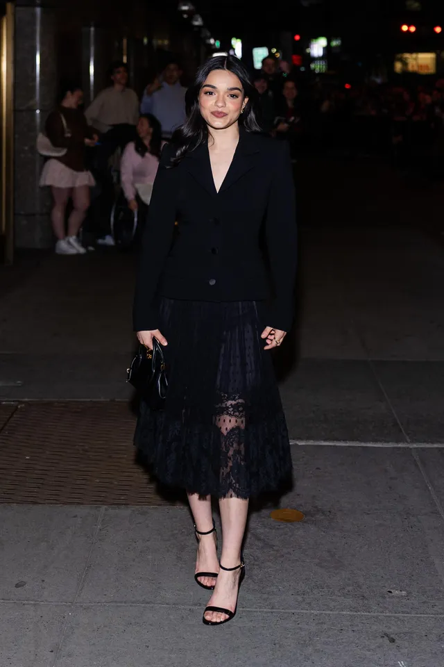 Rachel Zegler walking outside in a black lace dress and heels.
