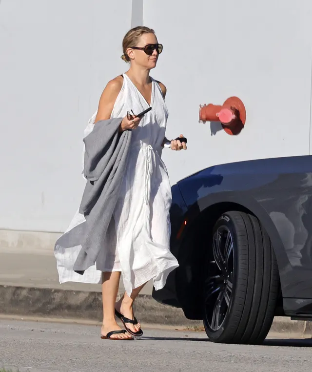 Kate Hudson walking beside a vehicle in a pale dress and flat sandals.