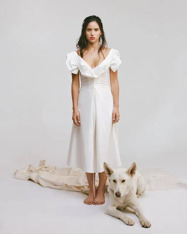 Adria Arjona standing barefoot in a white dress beside a dog.