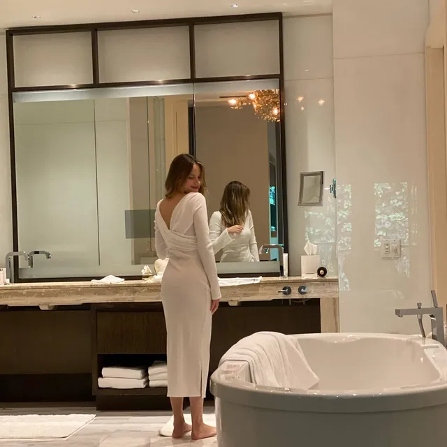 Halston Sage posing barefoot in a white robe beside a bathroom mirror.