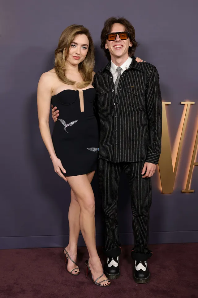 Peyton List posing beside a man in a black dress and heeled sandals.
