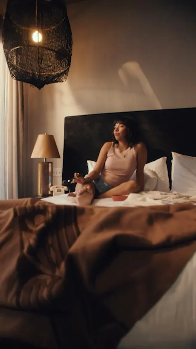 Xochitl Gomez sitting barefoot on a bed in a warm-lit room.