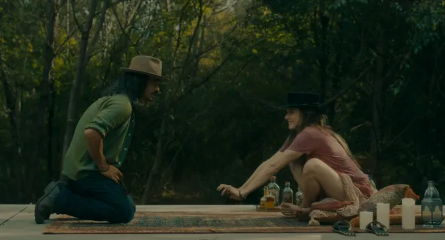 Rebecca Ferguson sitting barefoot on a wooden platform beside a companion in a wooded setting.
