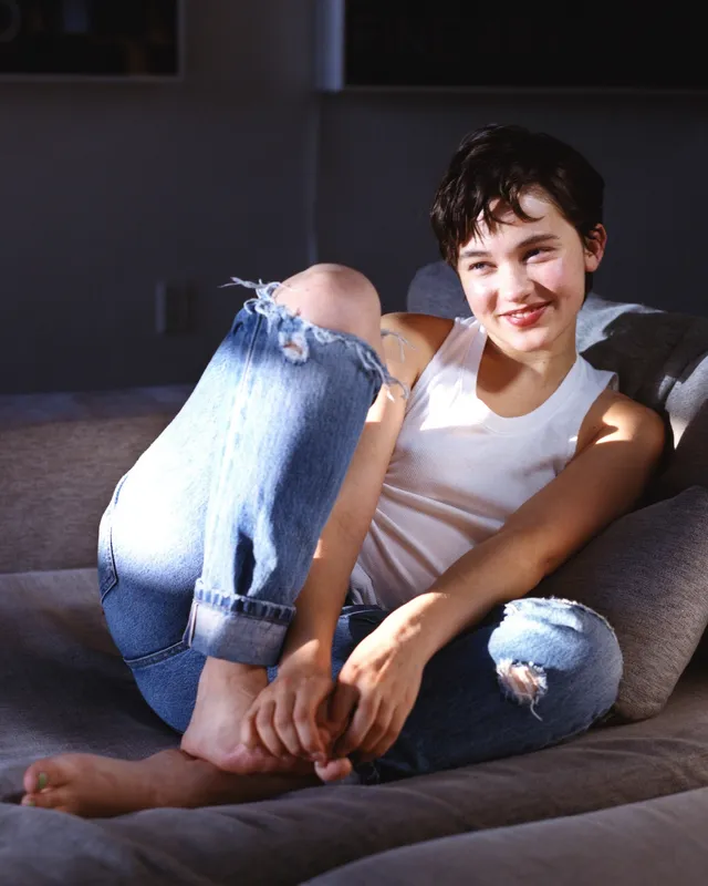 Cailee Spaeny sitting barefoot on a sofa in ripped jeans.