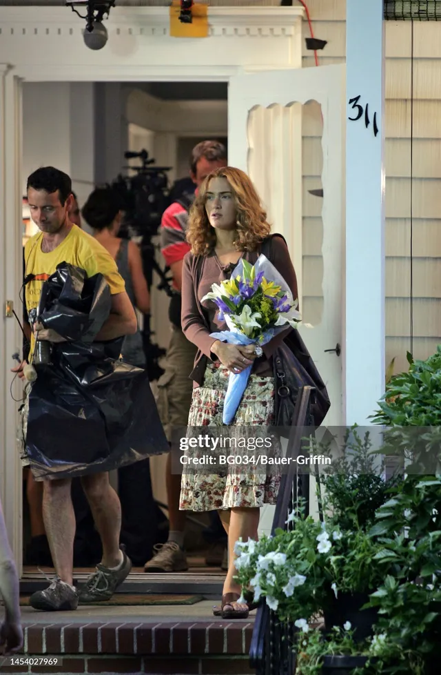 Kate Winslet standing outdoors in a floral skirt holding flowers.