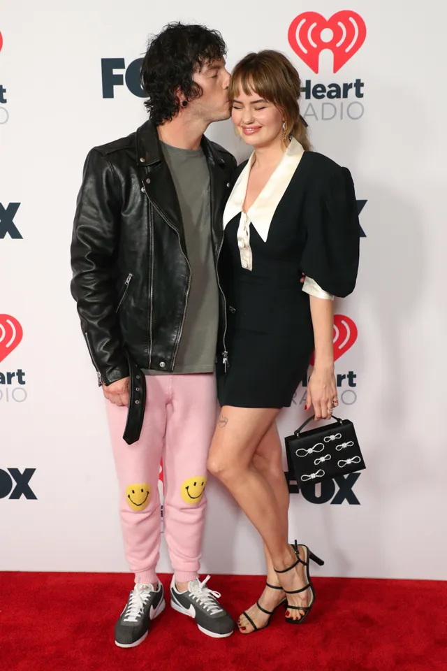 Debby Ryan posing with a guest in a black dress and heels on a red carpet.