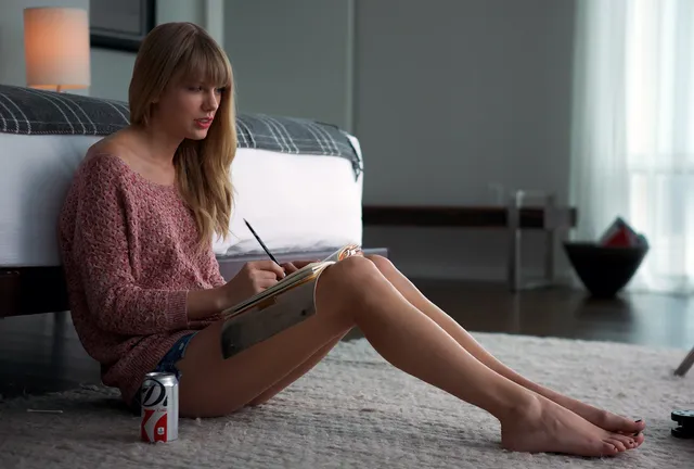 Taylor Swift sitting barefoot on a floor beside a sofa while writing.