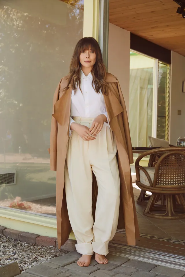 Elizabeth Olsen wearing a pale outfit and coat while standing barefoot.