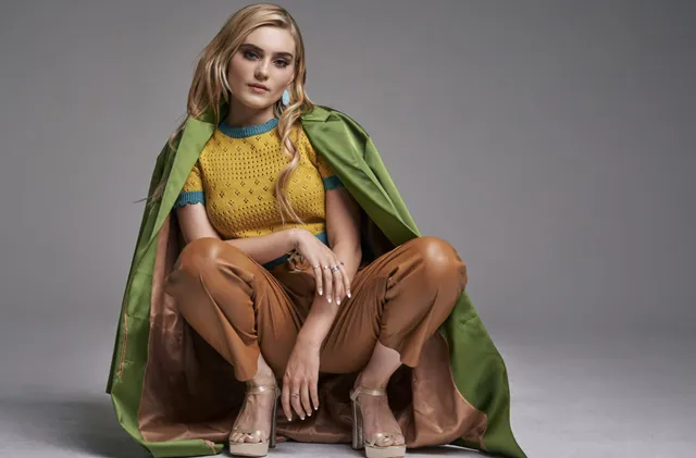 Meg Donnelly crouching in a colorful outfit and clear heels for a studio portrait.