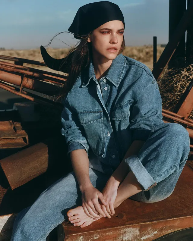 Barbara Palvin sitting barefoot in a denim outfit outdoors.