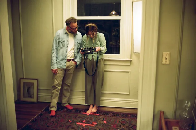 Olivia Wilde standing barefoot in a doorway beside another person in a film scene.