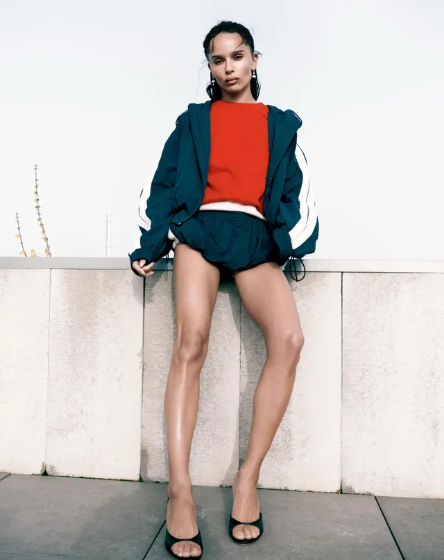 Zoë Kravitz posing in shorts and a jacket beside a wall.