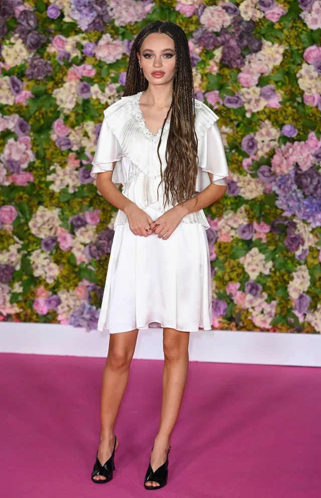 Nico Parker standing against a purple backdrop in a short white dress and heels.