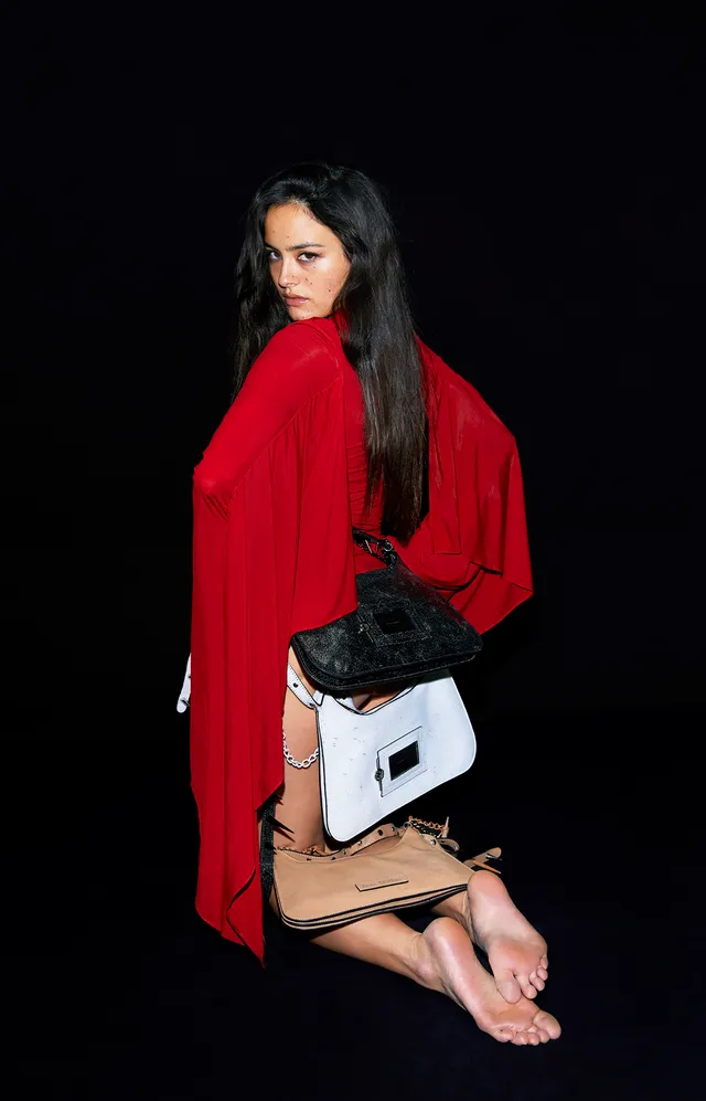 Chase Sui Wonders kneeling barefoot in a red outfit with handbags.