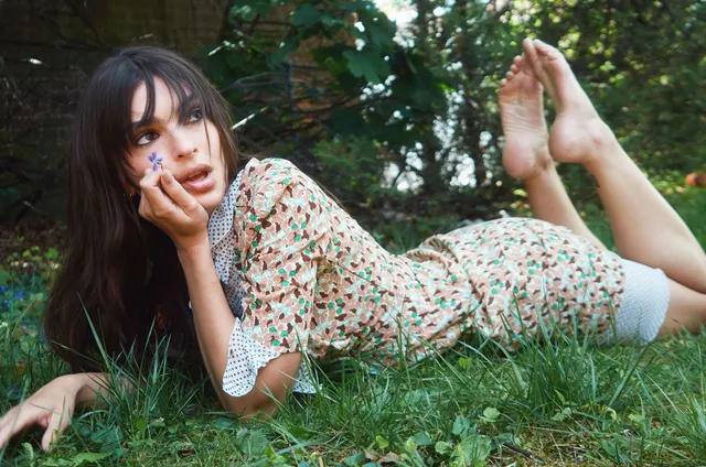 Emily Ratajkowski lying barefoot on grass in a patterned shirt.