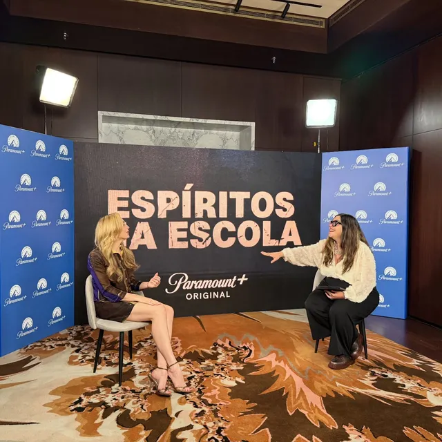Peyton List sitting during an interview in a dark dress and heels.