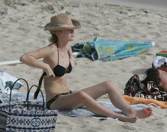 Kirsten Dunst sitting on a beach towel in a black swimsuit.
