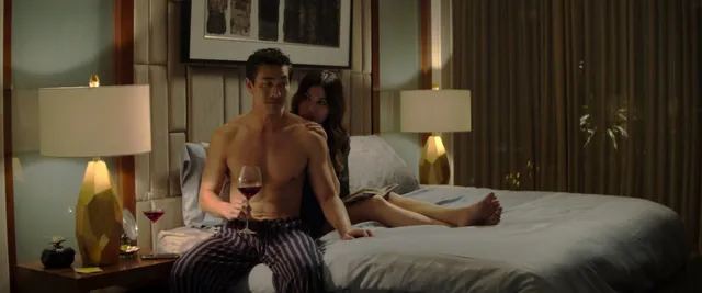 Gemma Chan sitting on a bed beside a man in a screen scene.