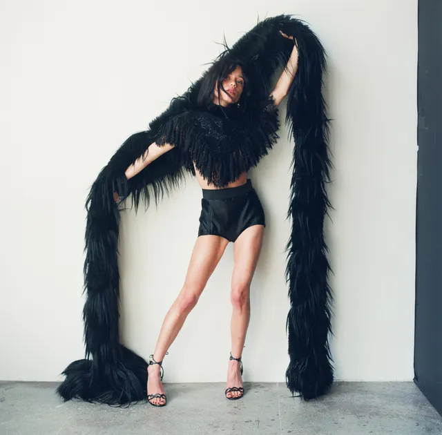 Jenna Ortega posing against a pale wall in a black outfit with feathered fabric and heels.