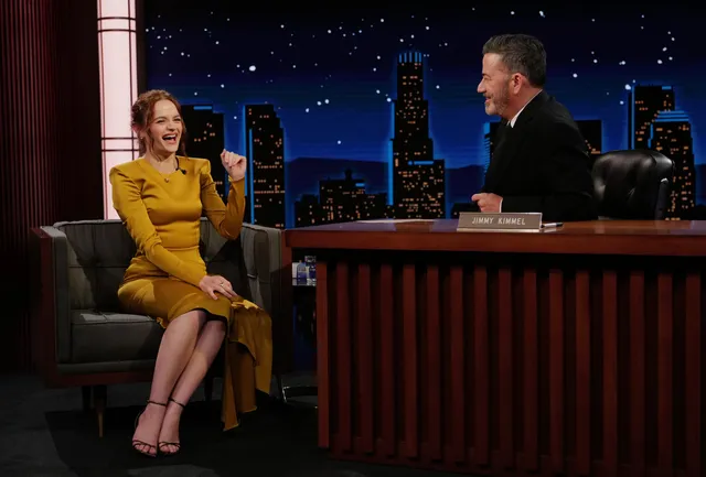 Joey King sitting for a television interview in a gold dress and heels.