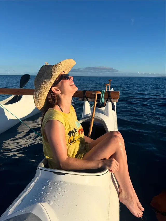 Emma Roberts sitting barefoot on a boat in a yellow shirt and straw hat.