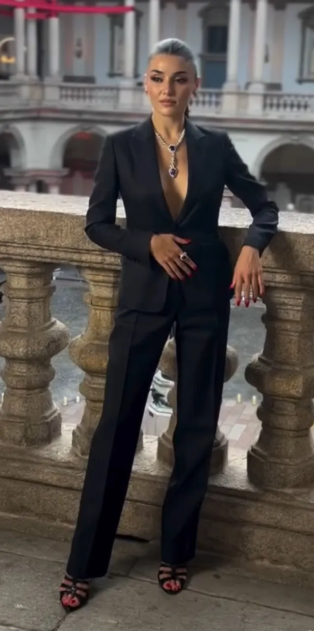 Hande Erçel standing by a balustrade in a black suit and heels.